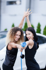 Two beautiful businesswomen in black outfut posing in front of an electric car. Girls holding blue charging cable. Shallow depth of field. Selective focus..
