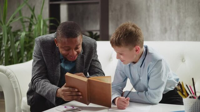 African-American man tutor laughs reading task in textbook with junior boy student at private English language lesson with native speaker slow motion