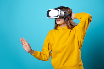 VR gamer, a young woman in a youth outfit uses a virtual reality helmet. Brunette in a yellow hoodie on a blue background with a virtual reality helmet on her head, full immersion