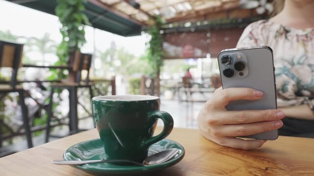 A girl in a sweater with flowers is sitting at a wooden table with a phone and a green cup of tea in a sunny cafe. A girl is resting on a warm summer day in a restaurant. Dolly shot. Slow motion.