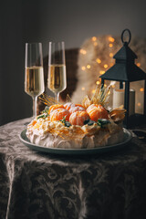 Pavlova cake is a meringue-based dessert named after the Russian ballerina decorated with fresh mandarins, caramel, mint, physalis, and powdered sugar with two champagne glasses on small table still.