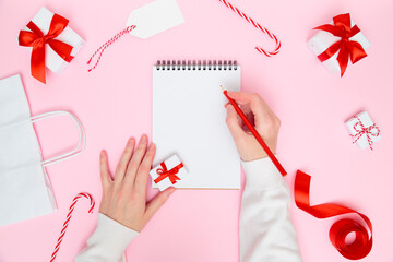 Christmas cozy background. Female's hand writing in blank notebook with festive decorations, gift boxes on pastel pink. New year planning, goals, to-do list or wish list concept. Flat lay, copy space.