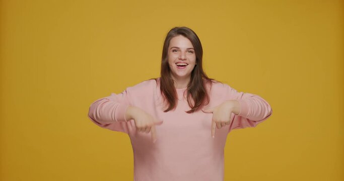 Joyful energetic young girl pointing down by fingers, recommend, advertise link, cool suggestion on yellow background