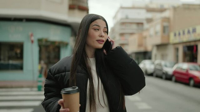 Young hispanic woman talking on the smartphone drinking coffee at street
