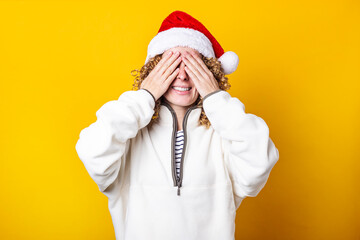 Smiling curly young woman in santa hat with palms closed her eyes on yellow background.