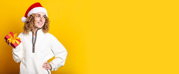 Smiling young woman in santa claus hat holding a gift on a yellow background. Banner.
