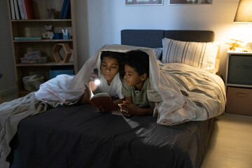 African American sister holding flashlight and reading book to brother while they lying in bed at night © pressmaster