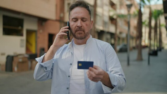 Young caucasian man talking on the smartphone holding credit card at street