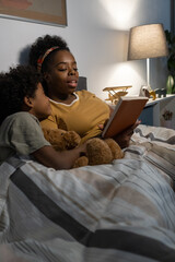 Careful African American blackwoman lying under duvet in bed and reading fairy tale to son