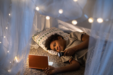 African American boy lying on pillow inside of tent and shining flashlight on book while reading...
