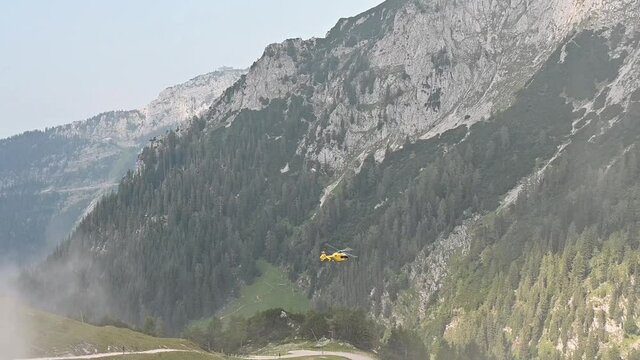 A yellow rescue helicopter flying away in front of an alpine panorama.