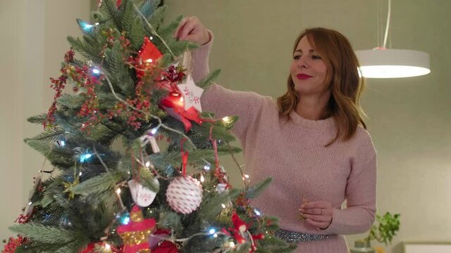 Woman decorating a Christmas tree in her living which is also decorated in the spirit of the upcoming holidays.