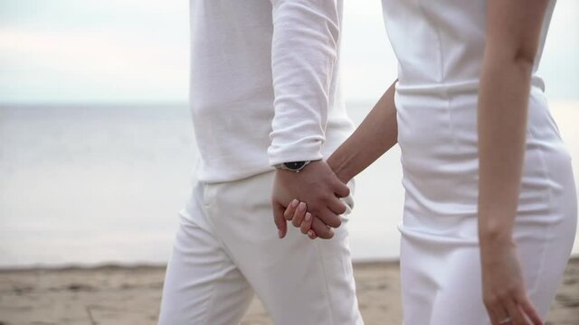 Young romantic couple in love walks at the beach in the evening sunset. Man and woman holding hands and hugs. Girl barefoot goes on sand. Bride and groom at their wedding in white dress and suit.