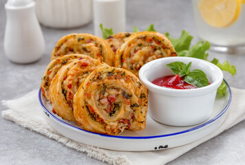 Baked puff pastry rolls or pinwheels with ham, cheese and herbs served on a plate with green salad and tomato dipping sauce. Party snacks idea. Close up, selective focus, gray concrete background.