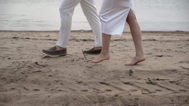Young romantic couple in love walks at the beach in the evening sunset. Man and woman holding hands and hugs. Girl barefoot goes on sand. Bride and groom at their wedding in white dress and suit.