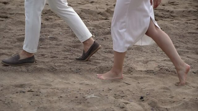 Young romantic couple in love walks at the beach in the evening sunset. Man and woman holding hands and hugs. Girl barefoot goes on sand. Bride and groom at their wedding in white dress and suit.