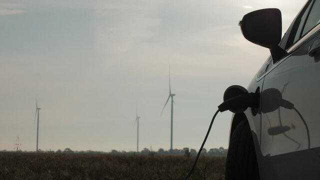 Wind turbines produce energy in field on cloudy day. White electric car charged by clean power stands against rotating windmills close view