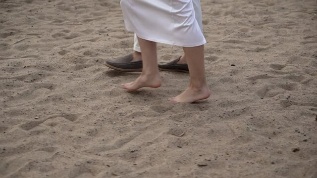 Young romantic couple in love walks at the beach in the evening sunset. Man and woman holding hands and hugs. Girl barefoot goes on sand. Bride and groom at their wedding in white dress and suit.