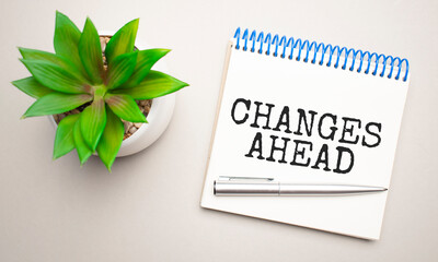 Changes Ahead is written in a white notepad near a clipboard, calculator, green plant, glasses and a pen on a yellow and concrete background. Business concept. Flat lay.