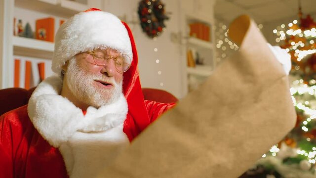 Santa Claus with a real white beard in a traditional red and white suit and round glasses is smiling sitting on a chair and holding a list of children's wishes