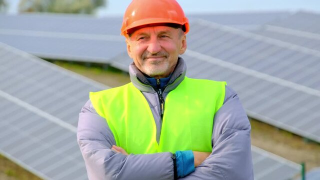 Solarcells installed at rural farm. Happy builder with beard in uniform crossed arms standing against sun panels at construction site closeup