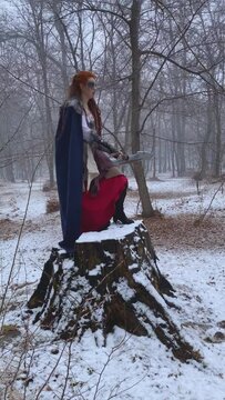 Male photographer explains to female model in Viking costume what position to stand in. Photo shoot for cosplayer in Celtic costume with axe in winter in snowy forest.