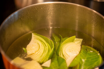 A top view of a stainless steel pot filled with boiling water, and cut pieces of organic cabbage cooking on a restaurant stove. The soup is being prepared for a meal.