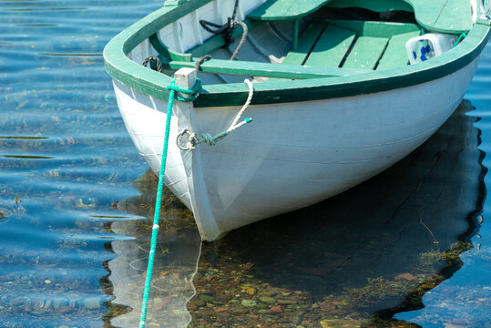 A white wooden traditional dory or small fishing vessel with green trim sits on a smooth water surface. The vintage dory has wooden oars and rope.  The water is reflecting the image of the boat.  
