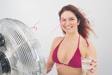 Red-haired smiling woman in a bikini drinks a cold drink and enjoys the blowing wind from an...