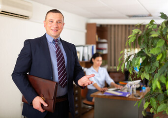 Fototapeta na wymiar Confident smiling man in business suit standing in modern office making inviting gesture with hand. Concept of invitation to business partnership