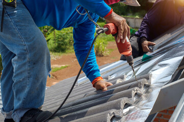 Roof repair, worker with white gloves replacing gray tiles or shingles on house with blue sky as background and copy space, Roofing - construction worker standing on a roof covering it with tiles.