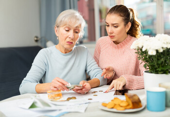 Frustrated young girl and elderly woman counting cash at table at home. Mother and daughter facing financials troubles .