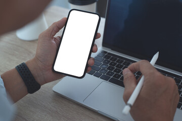 Mockup image of mobile phone, business man hand holding blank screen mobile phone during working on laptop computer at home office