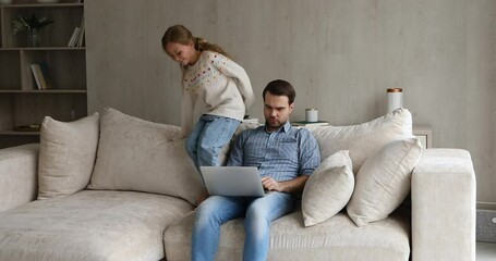 Serious dad work from home try to focus attention on pc screen read document online do business task while mischievous little girl jump on couch near. Small daughter behave bad disrupt job of father