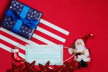 American flag composition with blue gift decorated with glittering stars, protective surgical mask and christmas tree decorations on red background. Space for text..