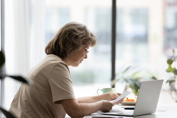 Focused senior homeowner, landlady reading paper document at laptop, doing domestic paperwork at home, paying paper bills, invoices, insurance fees online on banking app, sitting at table. Side view
