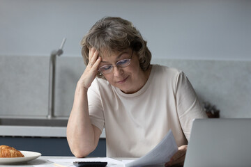 Concerned middle aged homeowner woman reading paper at laptop at home, reviewing bill for payment, thinking on financial document, insurance agreement, monthly fees, touching head