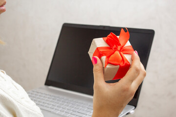 A woman's hand holds a gift box with a red bow and a laptop in her hands. The concept of online shopping for holiday gifts. Give gifts online via the Internet.