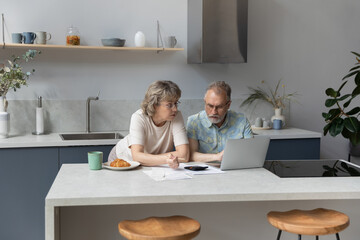 Serious older 60s married couple discussing budget, income, expenses, calculating money, counting insurance fees at calculator, doing paperwork together. paying bills, taxes, using laptop in kitchen