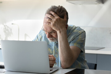 Frustrated tired senior man touching head at laptop, receiving shocking bad news, siffering from...