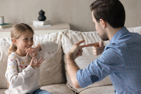 Happy adorable small kid girl with born hearing disability making gesture, using sign language communicating with caring young father or practicing with professional male Caucasian therapist.