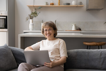 Happy middle aged lady holding laptop, sitting on comfortable couch, smiling, looking at camera. Senior pensioner woman using online app on computer, shopping on Internet. Home portrait