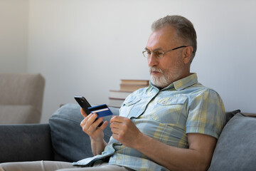 Focused senior older man using banking app, financial virtual service on mobile phone, paying bills, purchase online by credit card, making payments, orders on internet stores. Ecommerce concept