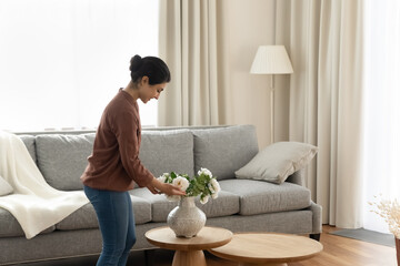 Smiling young indian ethnicity woman homeowner putting beautiful flowers in vase on coffee table in modern living room, enjoying decorating own apartment, improving styling interior on weekend. - Powered by Adobe