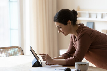 Beautiful smiling attractive millennial indian woman leaning over table, using digital tablet web...