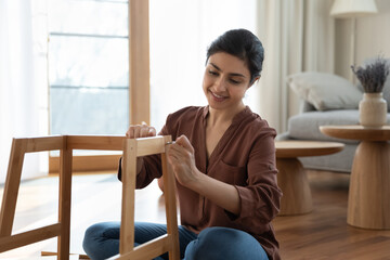 Happy beautiful young indian ethnic female homeowner fixing wooden chair or assembling new DIY...
