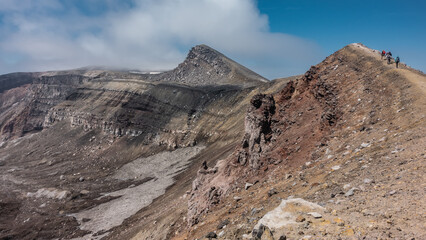 The trail runs along the edge of a volcanic crater. The layered structure of the soil. Tiny silhouettes of climbing tourists are visible on the path. Blue sky, clouds. Kamchatka. Gorely Volcano