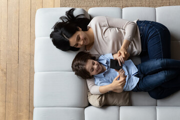 Woman and little 6s son lying on cozy sofa with smartphone, above view. Family spend leisure at home using modern tech usage, play games, buy goods, enjoy new cool amusing mobile app for kids concept