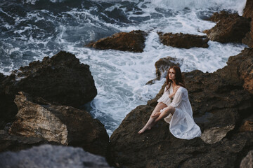 beautiful woman in a white wedding dress sits on the stones by the ocean pensive look