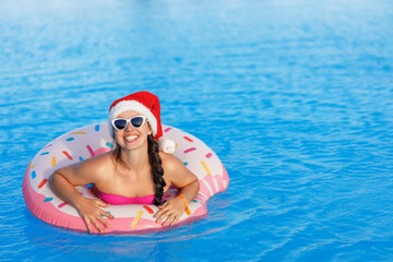 portrait of happy young woman in santa hat and sunglasses relaxing on inflatable ring in swimming pool